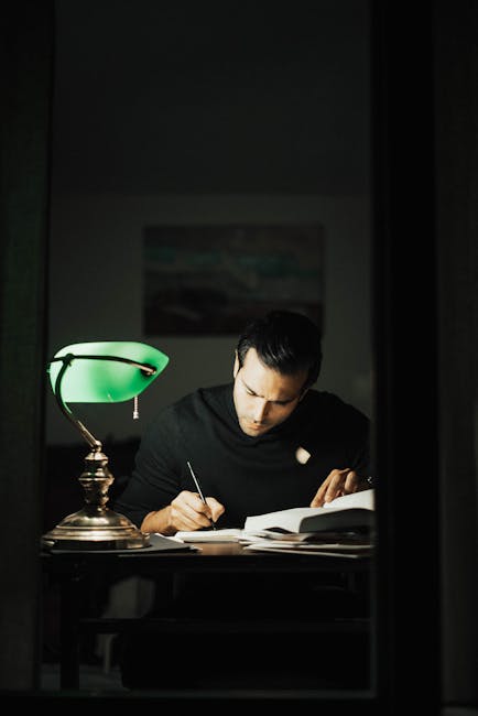 pexels-photo-7063764-7063764 Concentrated young male wearing black turtleneck taking notes and reading books while sitting at desk with papers and bankers lamp in dark home office